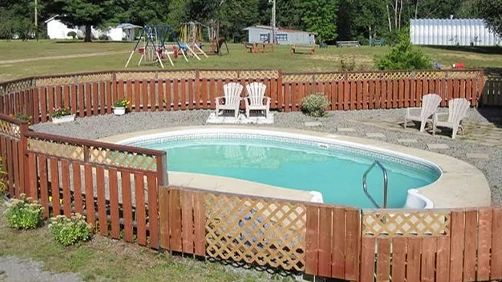 A Small, Oval Swimming Pool Is Surrounded By A Wooden Fence In A Backyard. Four Chairs Sit Nearby, And A Playground With Swings Is Visible In The Background.