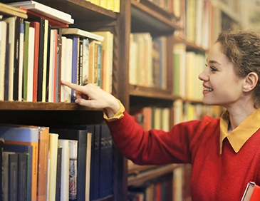 A Woman In A Red Sweater Smiles While Reaching For A Book On A Library Shelf. The Setting Is Warm And Inviting, Filled With Colorful Books.