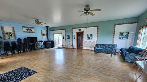 Spacious Living Room With Light Wood Floors, Blue Walls, And A Ceiling Fan. Features Blue Couches, Stacked Chairs, And Large Windows Creating An Airy Feel.